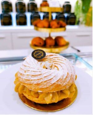 Bolo Paris Brest. Um bolo redondo com recheio cremoso e polvilhado com açúcar em pó está sobre um prato branco. Ao fundo, os bolos estão expostos em tabuleiros dourados e vêem-se prateleiras com frascos pretos.