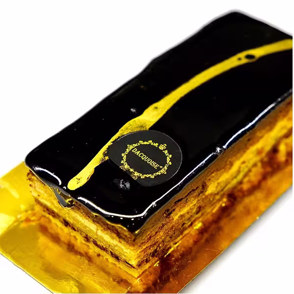 Um Bolo Opéra retangular com uma cobertura de chocolate preto brilhante, decorado com uma faixa diagonal dourada e um rótulo redondo, colocado elegantemente numa bandeja dourada.