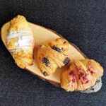 Três croissants com diferentes coberturas estão dispostos numa tábua de servir de madeira sobre um fundo escuro. Cada croissant francês aparece dourado e escamoso, decorado com açúcar, chocolate e pedaços de frutos silvestres.