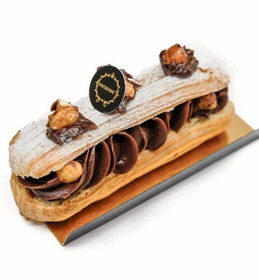 Um éclair recheado com um rico creme de chocolate, coberto com avelãs, decorações de chocolate, açúcar em pó e uma etiqueta redonda das nossas Coleções Sazonais, apresentado numa tábua retangular dourada sobre um fundo branco.