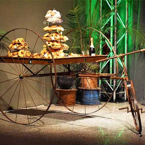 Um carrinho de madeira rústico com grandes rodas de metal exibe pilhas de bagels e pastelaria, juntamente com copos e uma garrafa de champanhe - perfeitos para o catering para festas particulares, com plantas verdes exuberantes e iluminação de palco.