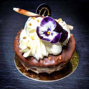 Uma sobremesa decadente dos bolos da Pâtisserie Dacquoise, coberta com chantilly, um pau de chocolate, granulado dourado e uma flor comestível roxa e branca numa base dourada, destaca-se como arte numa galeria contra um fundo escuro.