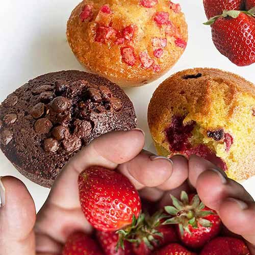 Três muffins - com pepitas de chocolate, morango e simples - são apresentados numa superfície branca. Em primeiro plano estão morangos frescos, com um toque doce.