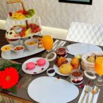 Uma mesa de pequeno-almoço posta para um brunch em Campo de Ourique, Lisboa, com pastelaria variada, macarons, fruta fresca, carnes frias, queijo, taças de cereais, sumo de laranja, um centro de mesa de flores vermelhas e duas cadeiras brancas ao fundo.