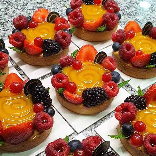 Pequenas tartes de fruta com recheio de creme amarelo brilhante, cobertas com morangos, framboesas, amoras, mirtilos, groselhas e folhas de hortelã.