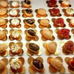 Um sortido de pequenos bolos decorativos dispostos em filas ordenadas sobre bases douradas, com vários toppings, incluindo chantilly, cobertura de chocolate e framboesas - perfeitos para catering ao domicílio ou festas particulares em Lisboa.