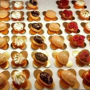 Um sortido de pequenos bolos decorativos dispostos em filas ordenadas sobre bases douradas, com vários toppings, incluindo chantilly, cobertura de chocolate e framboesas - perfeitos para catering ao domicílio ou festas particulares em Lisboa.