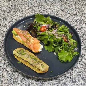 Um prato preto com uma salada verde mista, um éclair com salmão fumado e queijo creme e outro com uma pasta verde salgada, numa bancada salpicada.