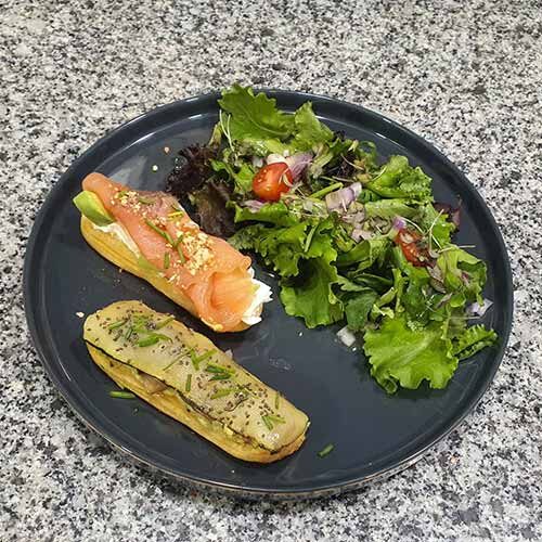 Um prato preto com uma salada verde mista, um éclair com salmão fumado e queijo creme e outro com uma pasta verde salgada, numa bancada salpicada.