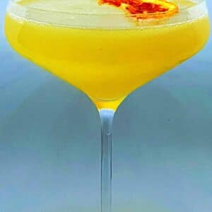 Um cocktail amarelo num copo coupé, decorado com uma pequena rodela de laranja seca, perfeito para festas particulares e eventos sofisticados, sobre um fundo claro e simples.