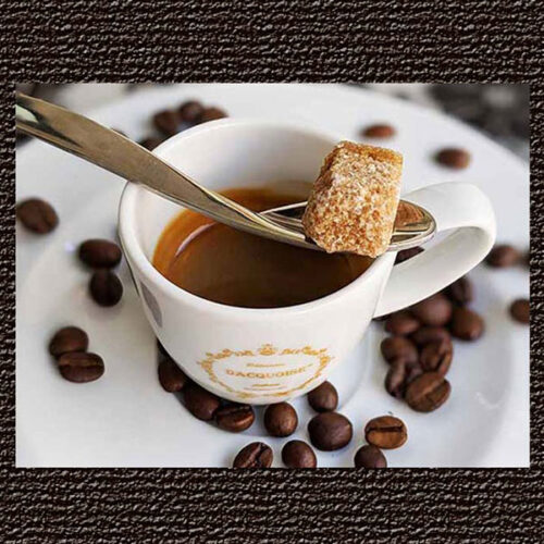 Uma chávena branca de café expresso assenta num pires com grãos de café espalhados. Uma colher equilibra-se na chávena, segurando um cubo de açúcar mascavado por cima do café, juntamente com sumos e chás.