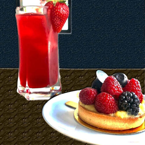Um copo de sumos vermelho decorado com um morango está ao lado de uma tarte de fruta coberta com framboesas, amoras e mirtilos num prato branco. O fundo escuro e texturado realça esta elegante apresentação do menu bebidas.