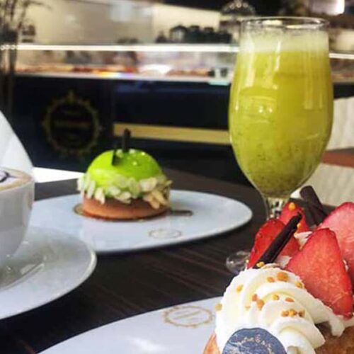 Uma mesa com sobremesas, incluindo uma tarte com morangos frescos e natas, uma massa verde brilhante, e um copo alto de sumos num ambiente acolhedor de pastelaria.