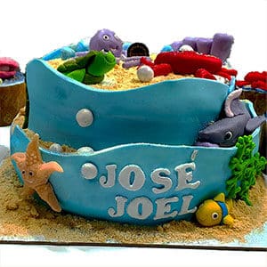 Um bolo azul com tema de oceano decorado com criaturas marinhas de fondant como uma tartaruga, tubarão, estrela do mar, peixe e caranguejo. O bolo tem toques de areia e o nome José Joel na frente-veja mais na galeria e fotos dos bolos da Pâtisserie Dacquoise.