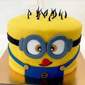 Um bolo redondo decorado à semelhança de um Minion banana, com cobertura amarela, óculos cinzentos, macacão azul e cabelo preto feito de cobertura no topo - apenas um dos bolos da Pâtisserie Dacquoise que encantam a nossa galeria.
