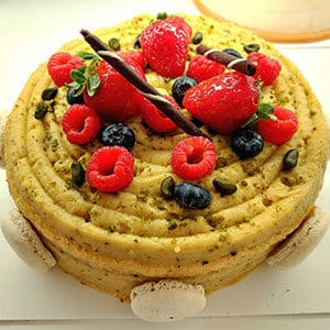 Um bolo redondo da nossa galeria, decorado com morangos, framboesas, mirtilos, sementes de abóbora, palitos de chocolate e rodeado de pequenos macarons brancos. A cobertura em redemoinho no topo é apresentada nas fotos dos bolos da Pâtisserie Dacquoise.