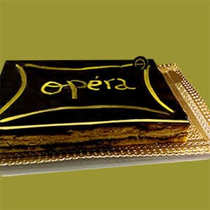 Um bolo de ópera retangular com cobertura de chocolate brilhante e a palavra opéra escrita a dourado, apresentado num tabuleiro dourado ornamentado sobre um fundo verde, dos bolos da Pâtisserie Dacquoise.