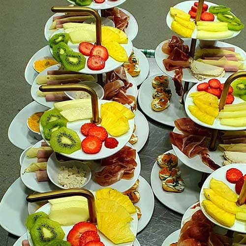 Uma exposição de tabuleiros dispostos em camadas com fatias de fruta fresca variada, incluindo kiwi, ananás, morangos e melão - perfeitos para um brunch vibrante em Campo de Ourique - juntamente com carnes enroladas e pequenos aperitivos em pratos brancos.