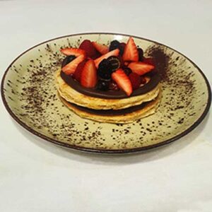 Uma pilha de panquecas cobertas com creme de chocolate, morangos fatiados e bagas variadas, servidas num prato polvilhado com cacau em pó - perfeito para catering para festas particulares ou catering para aniversários em Lisboa.