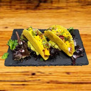 Dois tacos de casca dura recheados com alface, guacamole, manga cortada e cebola roxa em cubos, servidos num prato de ardósia preta com mistura de verduras , tudo muito bem apresentado numa superfície de madeira.