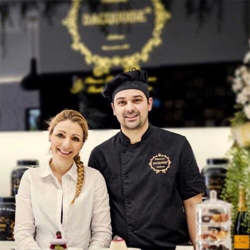 Uma mulher sorridente e o Chefe Ivo Camões estão atrás de um balcão de pastelaria com bolos e pastéis expostos em frascos de vidro. Um letreiro preto e dourado de uma pastelaria é visível ao fundo, dando um vislumbre de quem somos.
