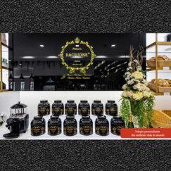 As prateleiras exibem latas pretas e douradas de chás sob um letreiro decorativo "Pâtisserie Dacquoise". Um ramo de flores brancas está ao lado, com cestos de produtos, uma máquina de café e um menu bebidas nas laterais.