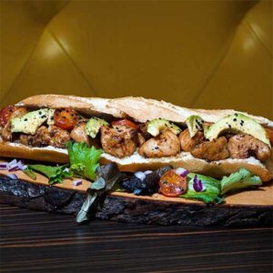 Uma sanduíche comprida com pedaços de frango grelhado, fatias de abacate, tomate cherry e folhas verdes, perfeita para catering para festas particulares, servida numa tábua de madeira rústica com salada e cebolas em cubos à parte.