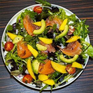 Uma salada colorida com verduras mistas, tomate cereja, salmão fumado, abacate e manga num prato branco sobre uma superfície de madeira escura.