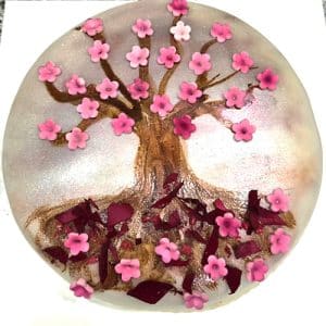 Um bolo redondo decorado com uma árvore feita de glacé castanho, coberto com muitas pequenas decorações de flores cor-de-rosa - perfeito para catering Lisboa ou catering e eventos. Pétalas e pedaços de flores cor-de-rosa estão espalhados na base da árvore.