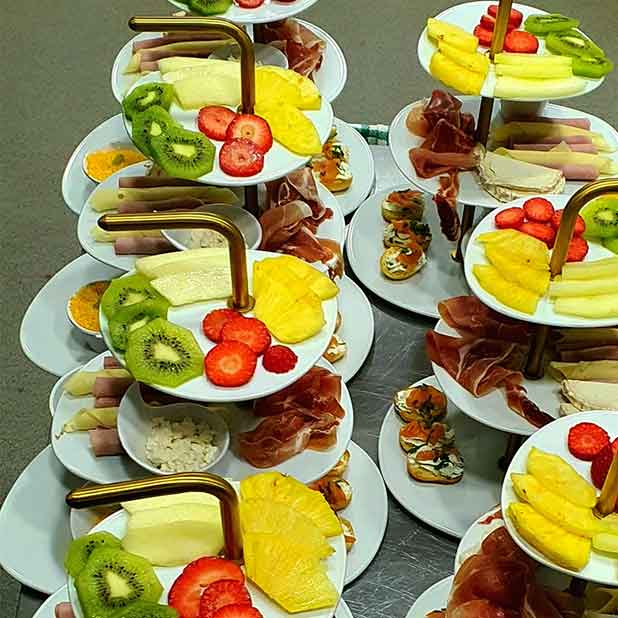 Uma exposição de tabuleiros dispostos em camadas com fatias de fruta fresca variada, incluindo kiwi, ananás, morangos e melão - perfeitos para um brunch vibrante em Campo de Ourique - juntamente com carnes enroladas e pequenos aperitivos em pratos brancos.