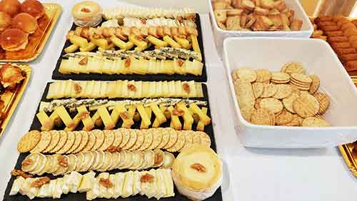 Uma mesa de buffet com queijos variados, bolachas, biscoitos e pãezinhos dispostos ordenadamente em tabuleiros e tábuas de servir pretas Alguns queijos estão decorados com nozes; alguns recipientes estão cobertos com película aderente.