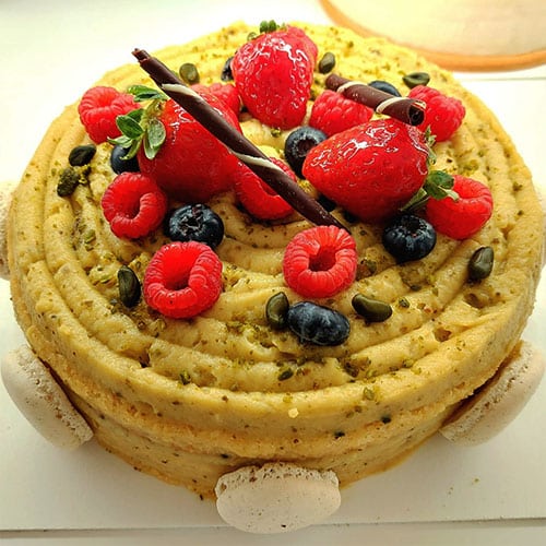 Um bolo redondo com um desenho em espiral, coberto com frutos vermelhos frescos, sementes de abóbora, palitos de chocolate e merengues. Delicie-se com esta criação da nossa linha de bolos sem glúten-ideal para quem procura bolos personalizados e deliciosos!.