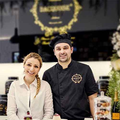 Uma mulher sorridente e o Chefe Ivo Camões estão atrás de um balcão de pastelaria com bolos e pastéis expostos em frascos de vidro. Um letreiro preto e dourado de uma pastelaria é visível ao fundo, dando um vislumbre de quem somos.