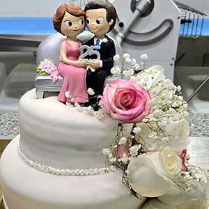 Bolo branco em camadas, decorado com rosas cor-de-rosa e brancas, encimado por uma estatueta de casal com um sinal de 25, perfeito para celebrar um 25º aniversário de casamento - ideal para catering ao domicílio ou catering para festas particulares.