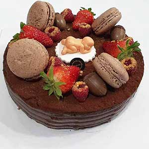 Bolo de chocolate decorado com macarons grandes, morangos, framboesas, pedaços de chocolate e uma pequena figura de bebé em fondant deitada sobre um disco branco no centro.