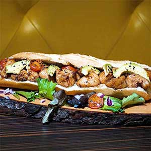 Uma sanduíche comprida com pedaços de frango grelhado, fatias de abacate, tomate cherry e folhas verdes, perfeita para catering para festas particulares, servida numa tábua de madeira rústica com salada e cebolas em cubos à parte.