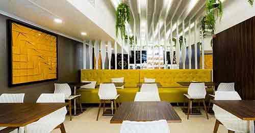 Interior moderno da Pâtisserie Dacquoise com bancos amarelos, cadeiras geométricas brancas, mesas de madeira escura, ripas verticais brancas, plantas verdes exuberantes e uma grande obra de arte abstrata amarela. Bem vindo a um ambiente convidativo da Pastelaria Francesa.