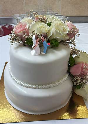 Um bolo branco dacquoise de dois andares decorado com rosas cor-de-rosa e brancas, encimado por uma estatueta de um casal, perfeito para celebrar um casamento com a elegância da Pastelaria Francesa.