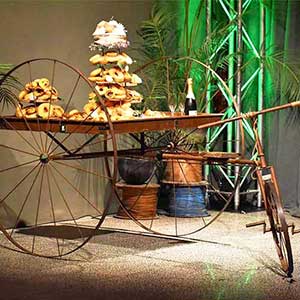 Um carrinho de madeira rústico com grandes rodas de metal exibe pilhas de bagels e pastelaria, juntamente com copos e uma garrafa de champanhe - perfeitos para o catering para festas particulares, com plantas verdes exuberantes e iluminação de palco.