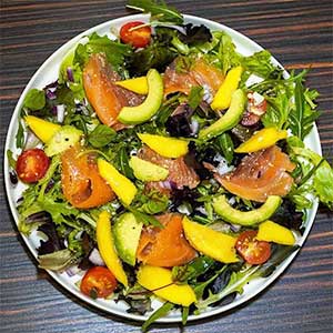 Uma salada colorida com verduras mistas, tomate cereja, salmão fumado, abacate e manga num prato branco sobre uma superfície de madeira escura.