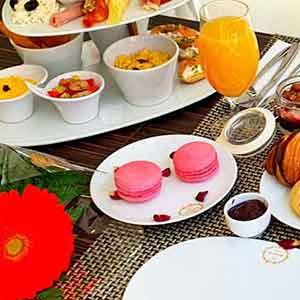 Uma mesa de pequeno-almoço posta para um brunch em Campo de Ourique, Lisboa, com pastelaria variada, macarons, fruta fresca, carnes frias, queijo, taças de cereais, sumo de laranja, um centro de mesa de flores vermelhas e duas cadeiras brancas ao fundo.