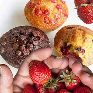 Três muffins - com pepitas de chocolate, morango e simples - são apresentados numa superfície branca. Em primeiro plano estão morangos frescos, com um toque doce.