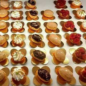 Um sortido de pequenos bolos decorativos dispostos em filas ordenadas sobre bases douradas, com vários toppings, incluindo chantilly, cobertura de chocolate e framboesas - perfeitos para catering ao domicílio ou festas particulares em Lisboa.