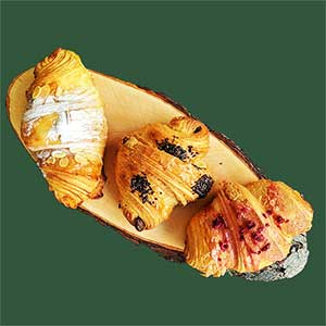 Três croissants variados, cada um com diferentes coberturas, estão dispostos numa tábua de servir de madeira rústica sobre um fundo verde liso, mostrando o toque artesanal da pastelaria fina.