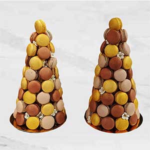Duas torres de macarons em forma de cone, adornadas com macarons amarelos, castanhos e beges e pequenas flores de fondant brancas, trazem a elegância da pastelaria fina a bases douradas sobre um fundo claro, tipo mármore.
