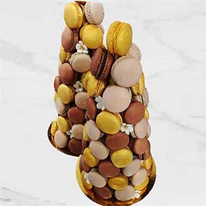 Duas torres de macarons em dourado, castanho e bege estão dispostas em bases douradas com delicadas flores brancas, perfeitas para catering Lisboa ou catering para festas particulares, sobre um fundo de mármore claro.