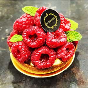 Uma tarte com framboesas frescas, folhas verdes de hortelã e um rótulo decorativo preto e dourado, assente numa superfície de textura escura - perfeita para os amantes da pastelaria fina e de tarteletes elegantes.