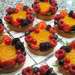 Pequenas tartes de fruta com recheio de creme amarelo brilhante, cobertas com morangos, framboesas, amoras, mirtilos, groselhas e folhas de hortelã.