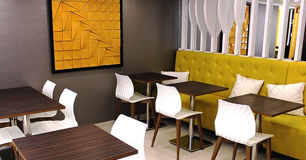 Interior moderno da Pâtisserie Dacquoise com bancos amarelos, cadeiras geométricas brancas, mesas de madeira escura, ripas verticais brancas, plantas verdes exuberantes e uma grande obra de arte abstrata amarela. Bem vindo a um ambiente convidativo da Pastelaria Francesa.