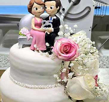 Um bolo branco em camadas, cor-de-rosa e brancas e encimado por uma estatueta de casal com um sinal de 25, a celebrar as suas bodas de prata.